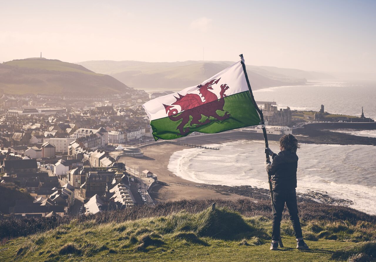 A child, standing on top of a hill, holding a Welsh flag looking over the coastline.