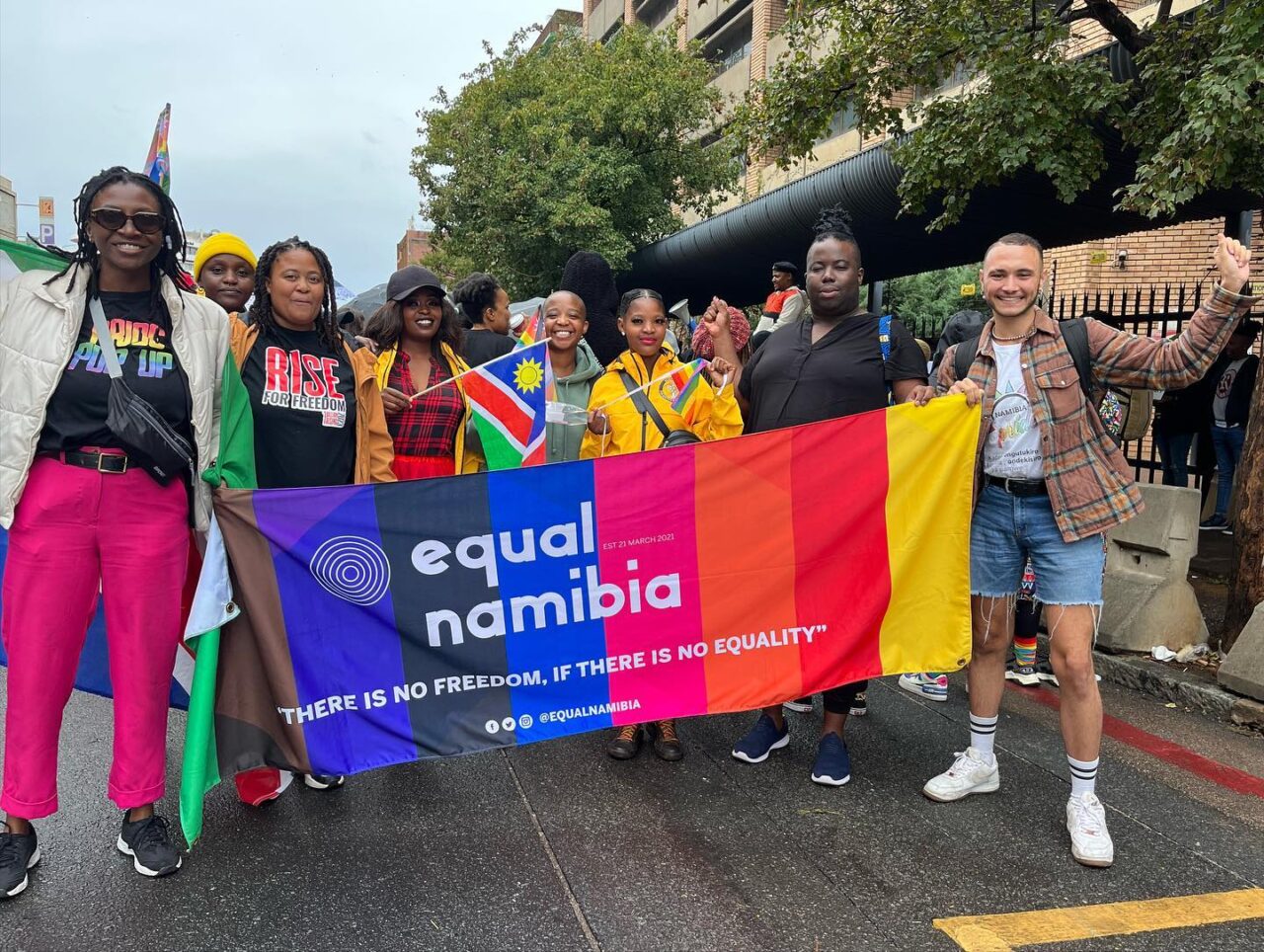 A diverse group of people holds a vibrant "Equal Namibia" banner at a pride event, displaying joy and unity. The banner reads, "Equal Namibia: There is no freedom, if there is no equality."
