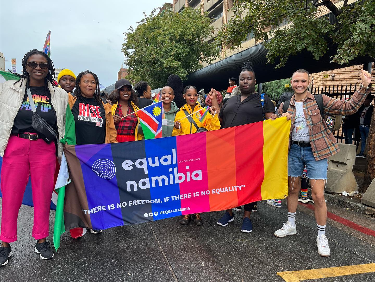 A diverse group of people holds a vibrant "Equal Namibia" banner at a pride event, displaying joy and unity. The banner reads, "Equal Namibia: There is no freedom, if there is no equality."